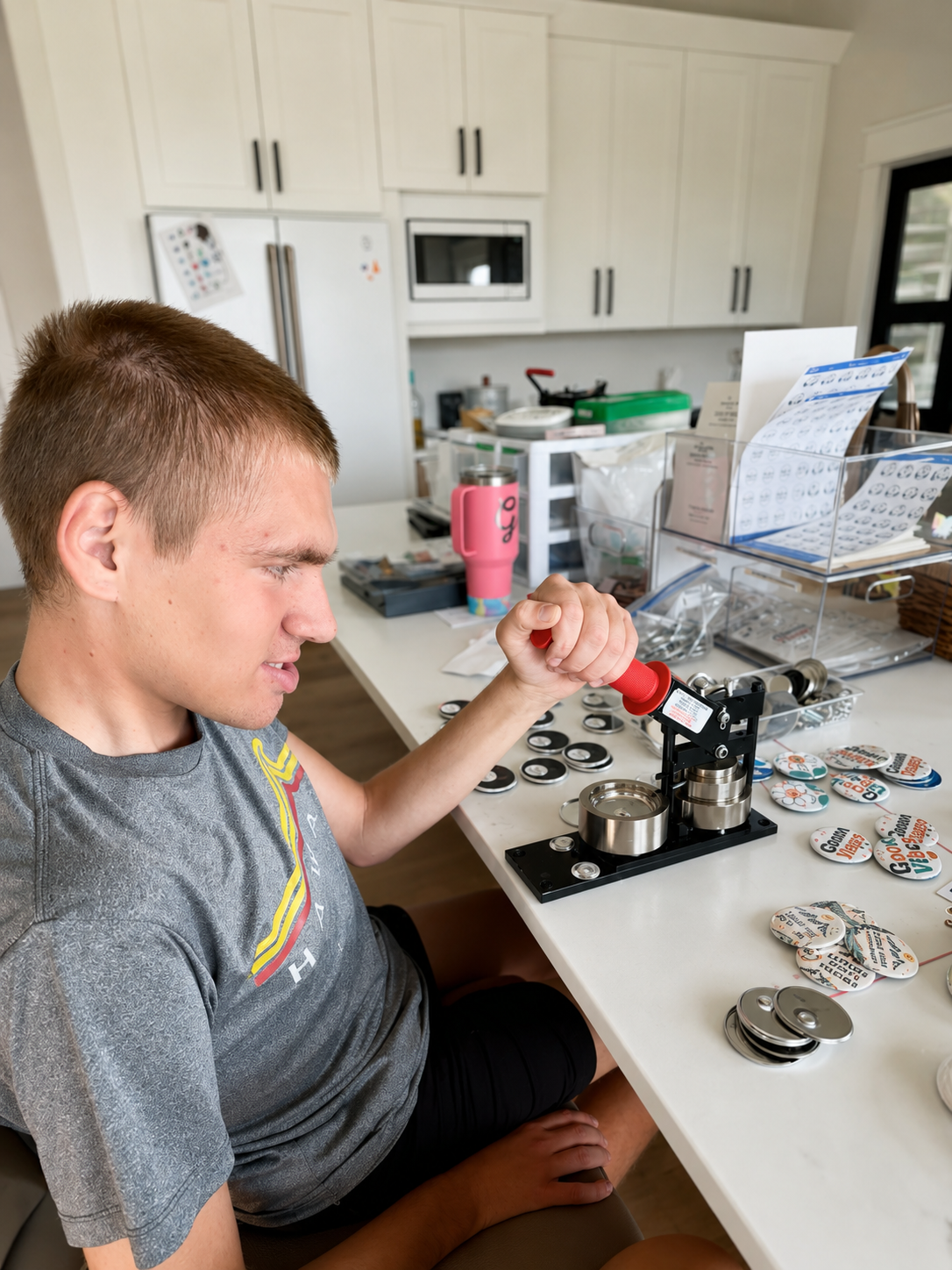 Carter uses a button maker at a worktable surrounded by finished Carter & Crew buttons.