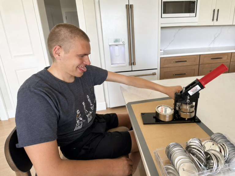 Carter smiles while using a button press to create Carter & Crew products at his worktable.